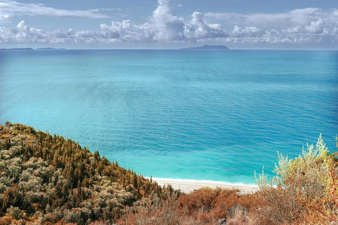 Blick von einem mit Macchia bewachsenen Hang auf türkisfarbenes Meer, einen hellen Strand und eine Insel am Horizont