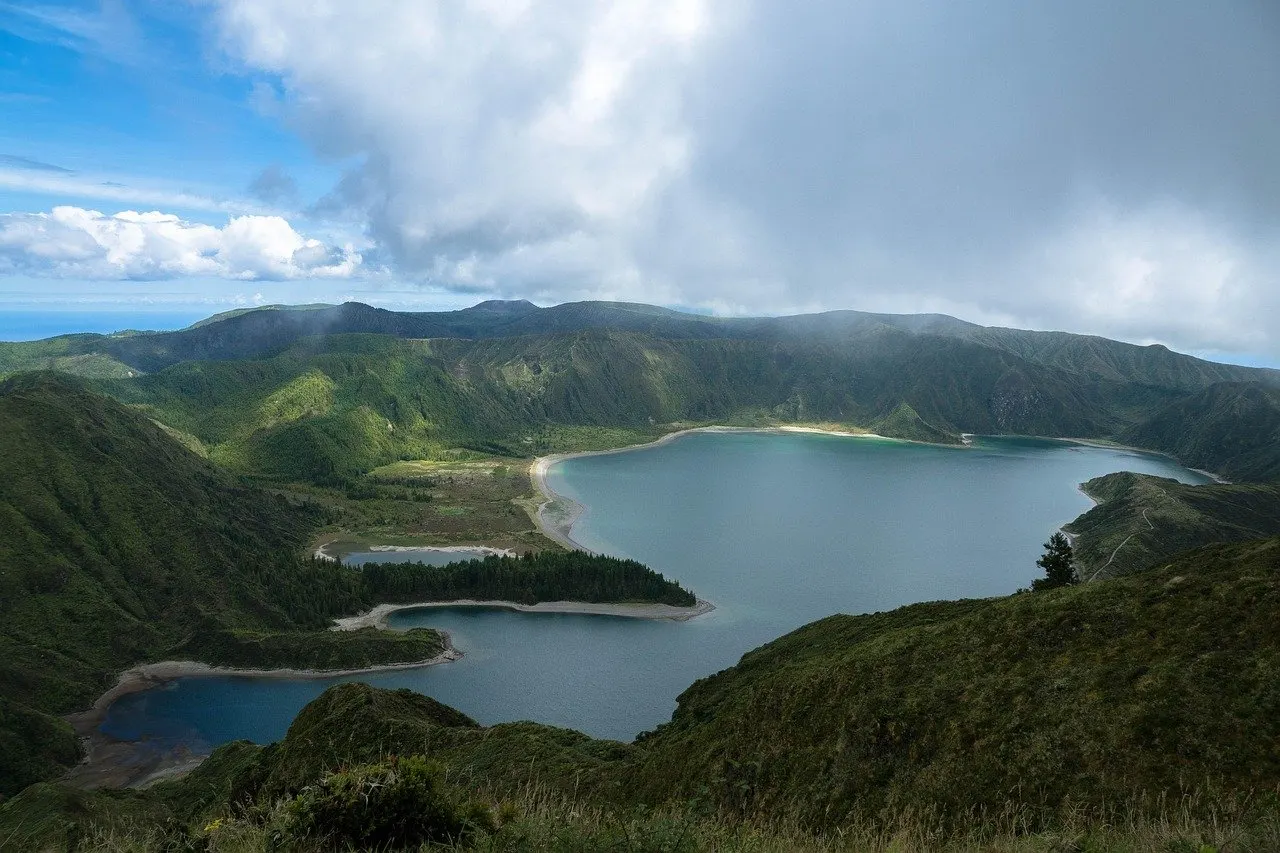 Kratersee mit hellen Sandufern zwischen grün bewachsenen Bergrücken auf den Azoren, darüber dichte Wolken und ein Stück blauer Himmel