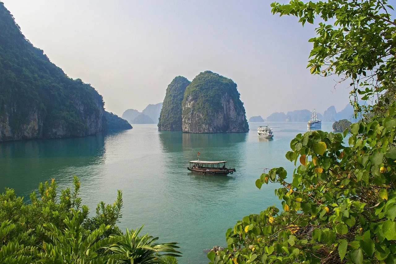 Bewachsene Kalkfelsen in der Ha-Long-Bucht, auf dem grünen Wasser ein kleines Holzboot und zwei weiße Ausflugsschiffe