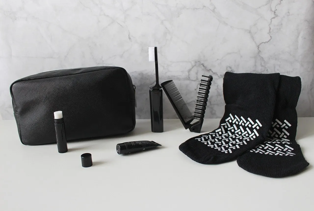 Schwarzer Kulturbeutel mit kleiner Cremetube, Lippenpflegestift, Reisezahnbürste, Klappkamm und Socken auf hellem Tisch