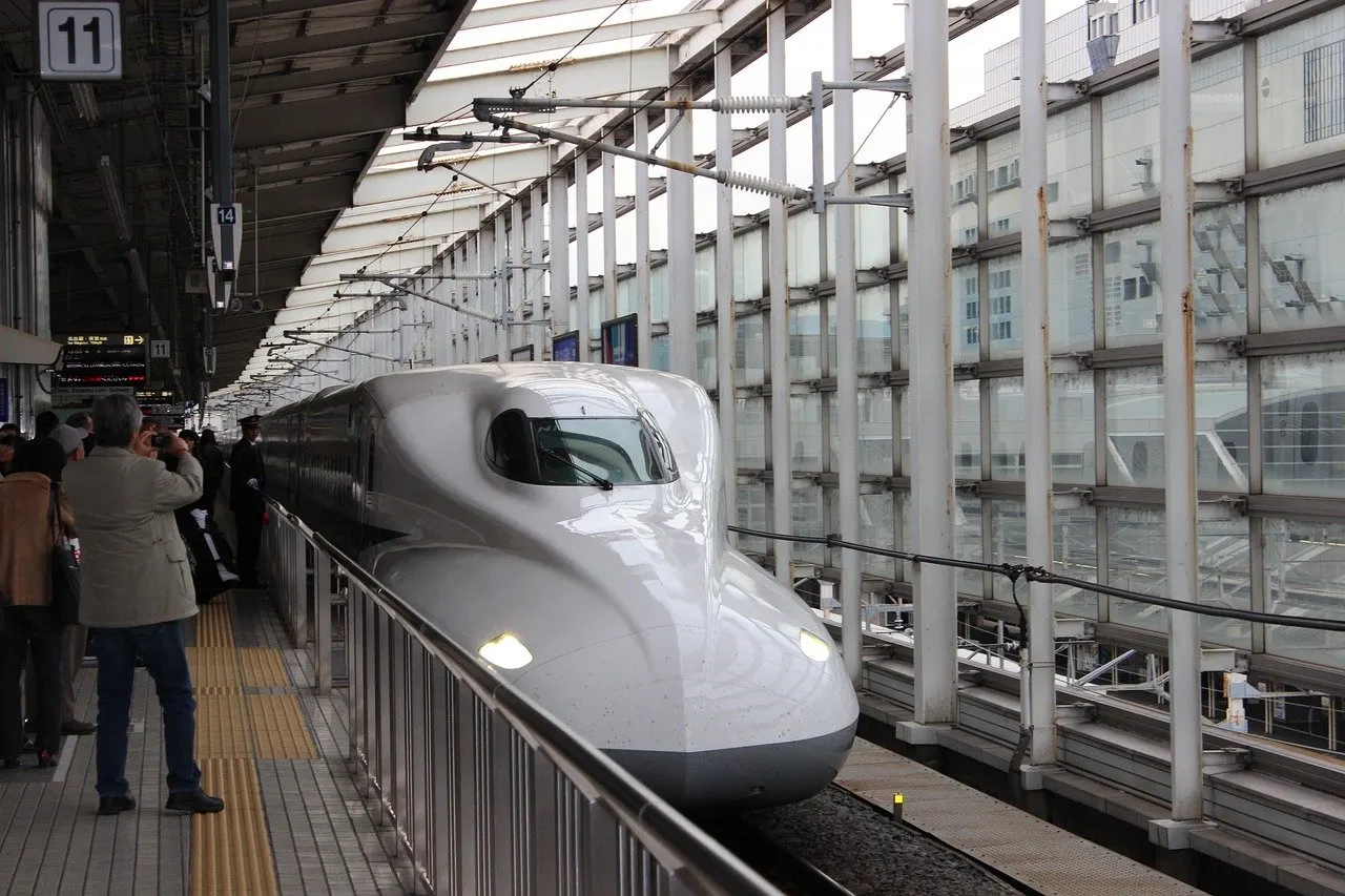 Weißer Shinkansen-Hochgeschwindigkeitszug der Baureihe N700 an einem überdachten Bahnsteig, im Hintergrund wartende Reisende