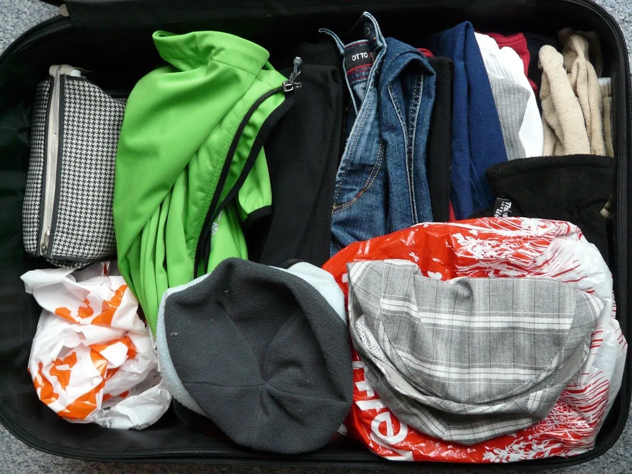Geöffneter schwarzer Koffer von oben, gefüllt mit Jeans, grüner Jacke, Mützen, Kulturbeutel und mehreren Plastiktüten