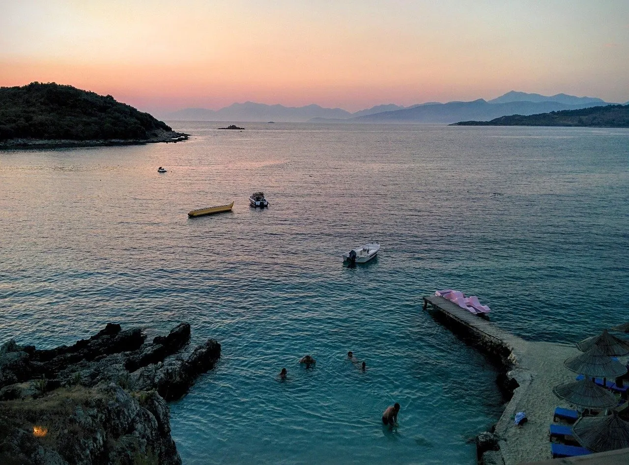 Abendstimmung in einer Bucht bei Ksamil mit Badestelle, Steg, Sonnenschirmen, kleinen Booten und Bergen am Horizont