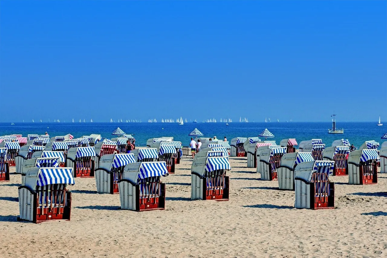 Reihen blau-weiß gestreifter Strandkörbe auf hellem Sandstrand an der Ostsee, dahinter blaues Wasser mit vielen Segelbooten
