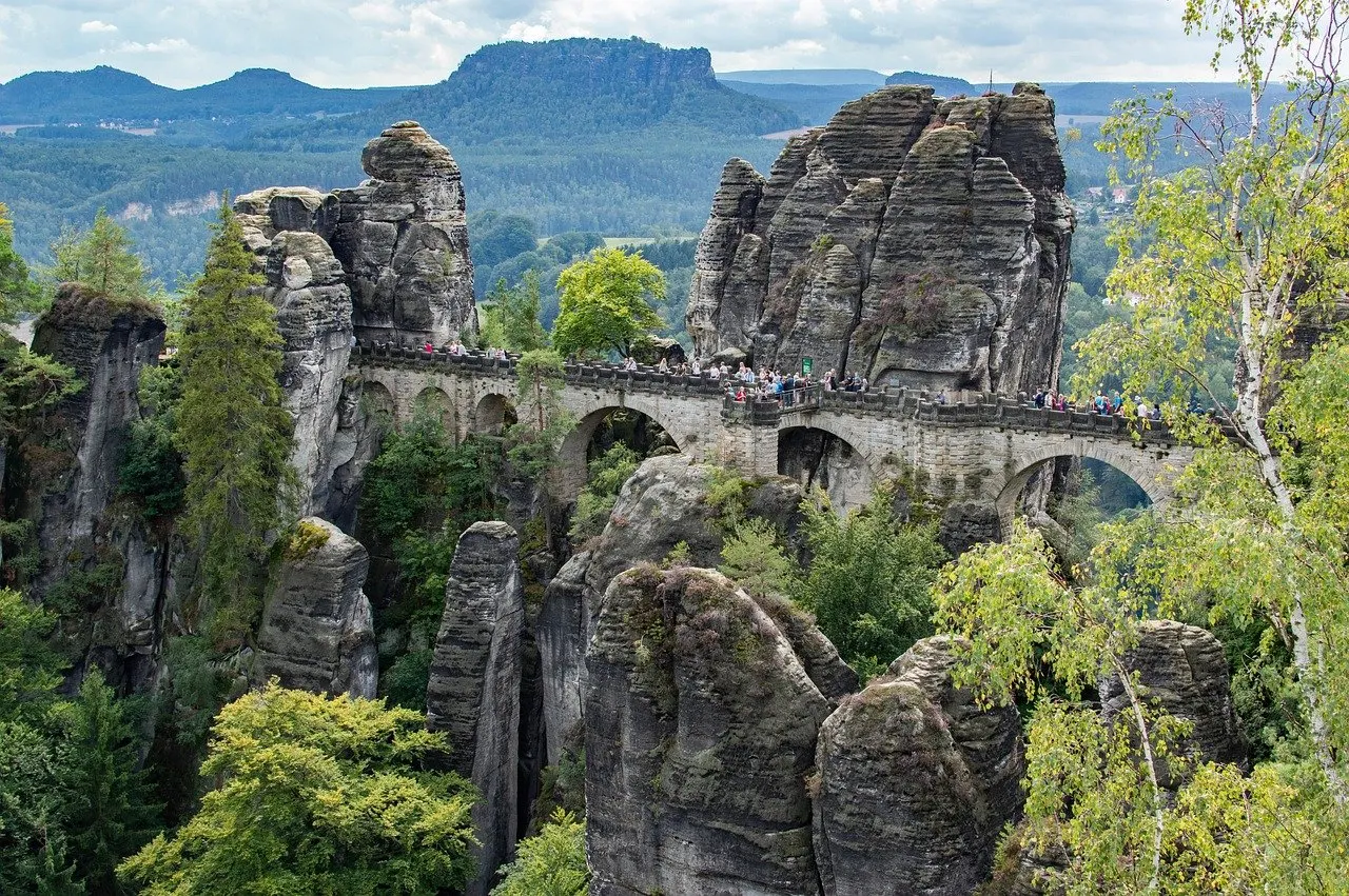 Steinerne Bogenbrücke mit Besuchern zwischen hohen Sandsteinfelsen an der Bastei, ringsum Laub- und Nadelbäume, dahinter Tafelberge unter leicht bewölktem Himmel