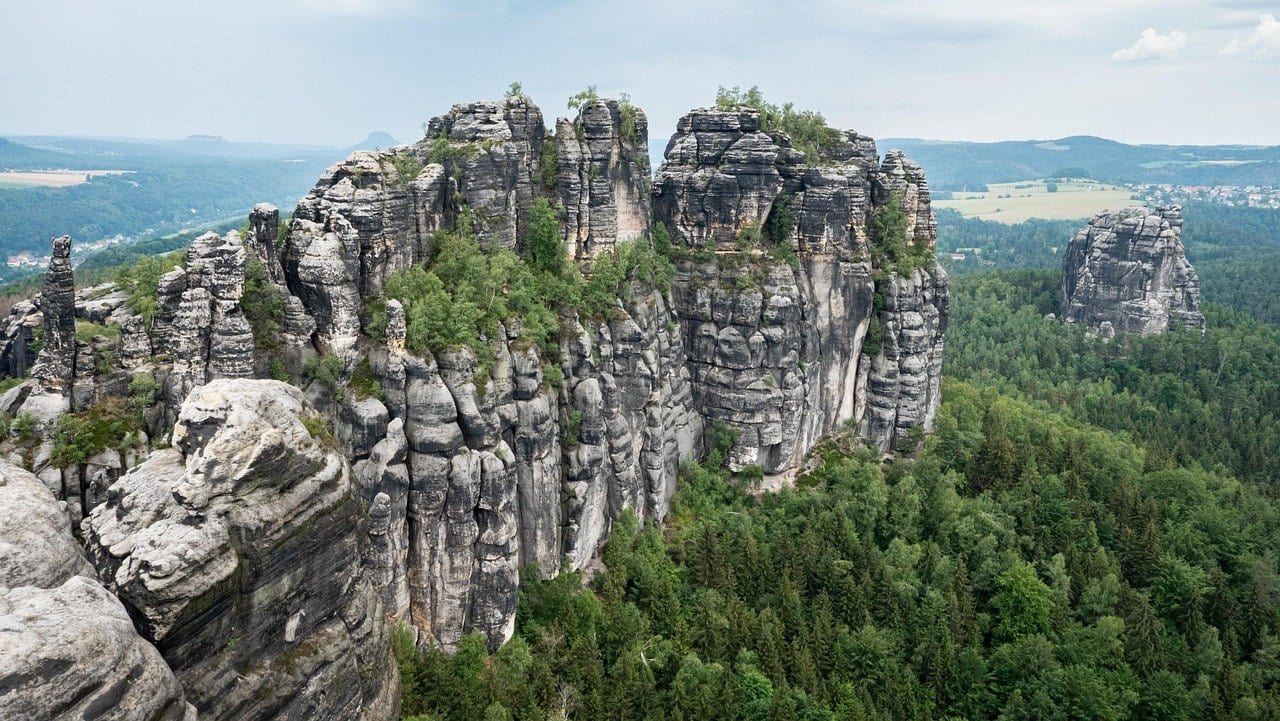 Zerklüftete Sandsteinfelsen der Schrammsteine mit vielen Türmen und Spalten, darunter dichter Nadelwald, im Hintergrund Hügelland
