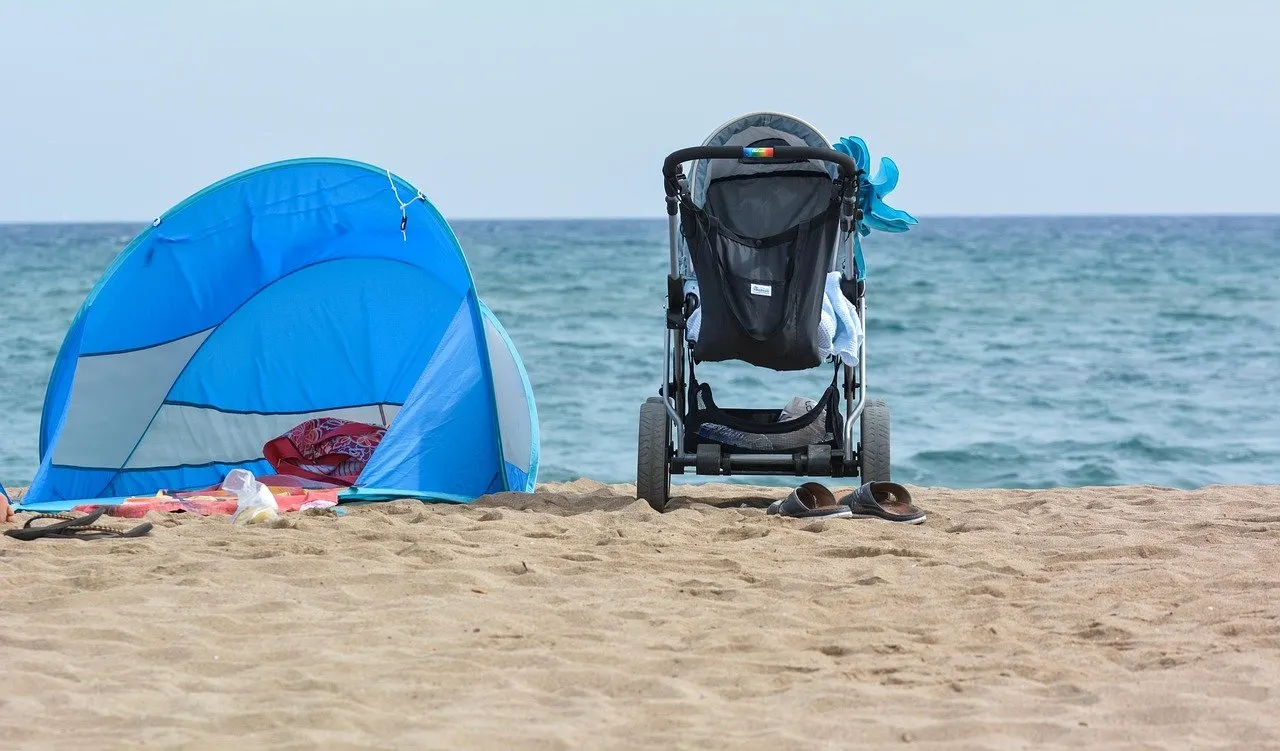 Kinderwagen und blaue Strandmuschel am Sandstrand vor dem Meer, vor dem Kinderwagen ein Paar Badelatschen